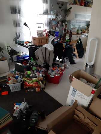 Unordentliche Küche und Wohnzimmer mit Kartons, Haushaltsgeräten, Spielzeug und Verpackungen.